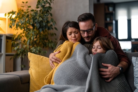 A family huddles together on a couch, wrapped in blankets, appearing distraught.