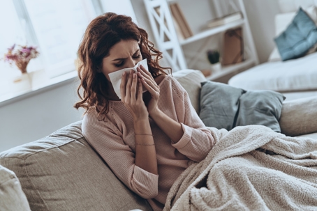 A woman is seen on a couch, bundled in a blanket, holding a tissue to her nose.
